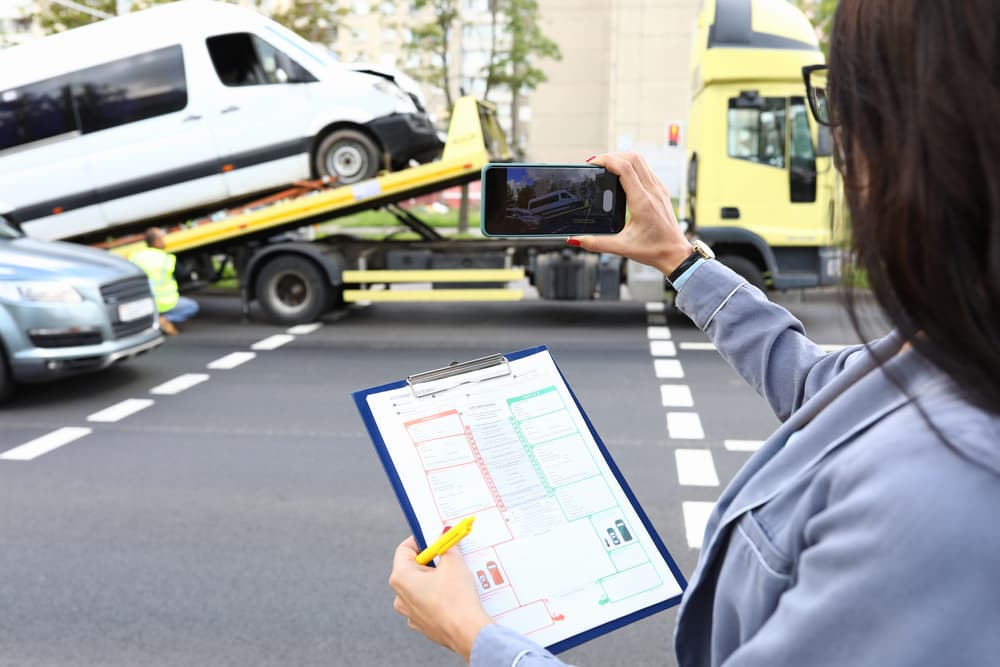 Insurance adjuster documenting a vehicle accident scene with a clipboard and smartphone as a damaged van is towed, representing multiple insurance companies involved.