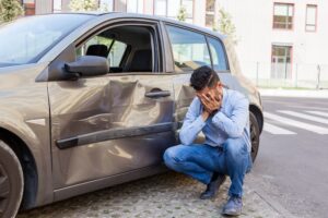 Distraught man holding head in hands next to a car with side damage.