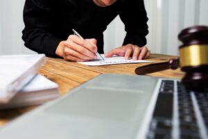 Person writing legal documents at a desk with a gavel and laptop, depicting the initial consultation with an attorney.