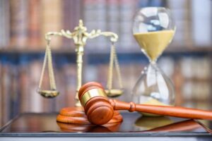 Symbolizing justice: Judge's gavel on a courtroom desk with blurred scales, bookshelf, and hourglass in a legal office setting.