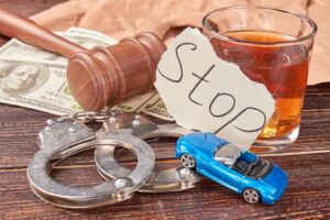 A judge's gavel, handcuffs, money, a glass of whiskey, a torn 'Stop' sign, and a miniature car displayed together.