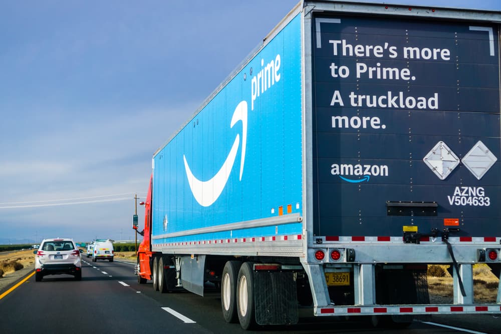 A blue Amazon Prime delivery truck on the road with promotional text, driving amidst other vehicles under a clear sky.