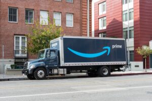 An Amazon Prime delivery truck parked on a city street beside multi-story buildings on an overcast day.
