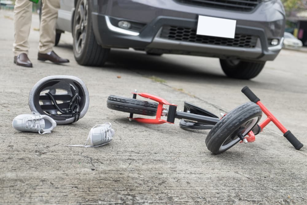 A child's bicycle lies wrecked near a car with helmet and shoes scattered, suggesting a fatal accident.