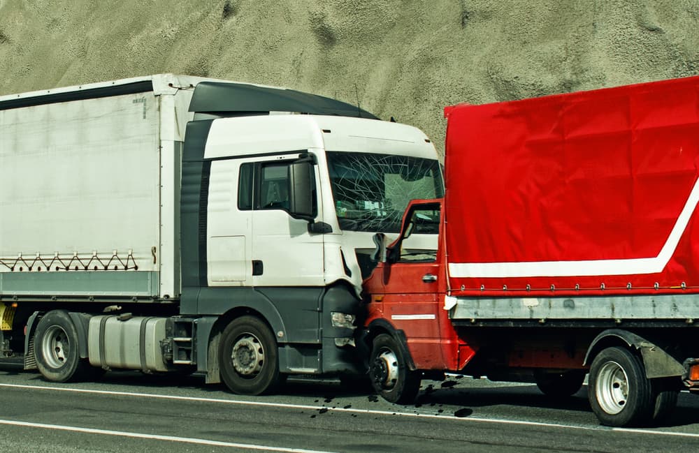 Collision between a white and a red truck on the roadside, with visible damage to the red truck's cab.