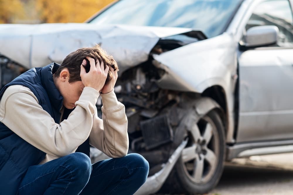 A distressed man beside a damaged car in Austin TX, holding his head in realization. The severity of the damage sinks in – the car is irreparable.