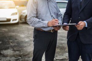 Car insurance agent offers pen to customer signing insurance form on clipboard while inspecting car after accident claim.
