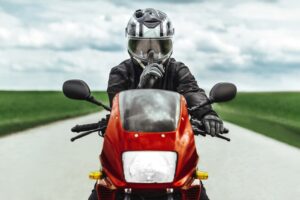 A man in protective gear and helmet sits on a red motorcycle, gesturing quietly with his finger. The biker knows a secret.
