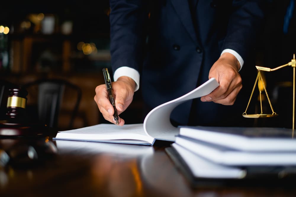 Lawyer signing documents at desk with a gavel and scales of justice in the background.