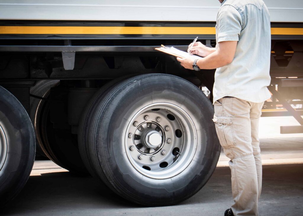 Vehicle inspector checking a commercial truck tire and suspension while taking notes on a clipboard during a safety inspection